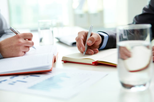 Two people sit at a desk, diligently taking notes. One person holds a pen over lined paper, while the other writes in a spiral notebook. A printed report discussing the burden of proof rests next to a glass of water in the bright office setting.