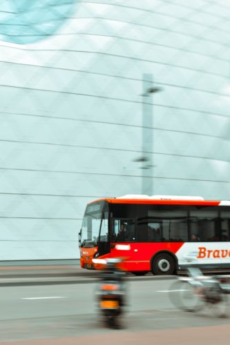 A red and white bus labeled "Bravo" moves swiftly down a Washington D.C. road. In the foreground, a motorbike and bicycle pass by, set against the backdrop of a modern geometric building facade, hinting at the intricate dance of urban mass transit.