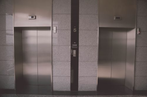 Two modern stainless steel elevators with closed doors are set in a gray marble wall, reminiscent of the sleek architecture often found in Washington D.C. A digital display above each door shows the floor number, and a glowing "5" is visible in the center between the elevators.