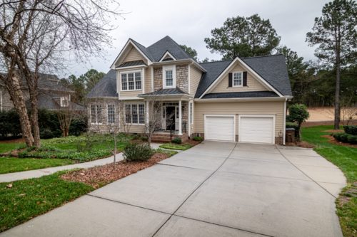 A two-story suburban house with gray siding and a gabled roof features large windows, a covered entryway, and a double garage. The front yard has landscaped bushes and a concrete driveway leading to the garage—ideal for meeting your personal injury attorney if you've been hurt at his house.