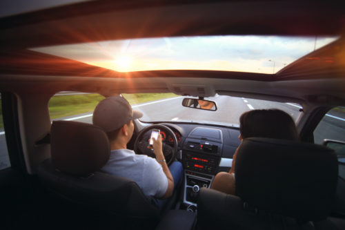 A man is driving on a highway near Washington D.C. at sunset, viewed from the backseat. He wears a cap and is texting while driving, as a passenger sits beside him. The panoramic sunroof reveals the sky and setting sun, creating a picturesque yet risky moment.