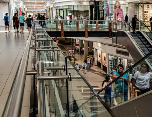 A bustling shopping mall in Washington D.C. features crowds navigating the upper and lower levels. On the right, an escalator is visible. Various storefronts, including a clothing store with large posters, line the corridor while glass railings divide the levels—watch for retail store injury hazards as you shop.